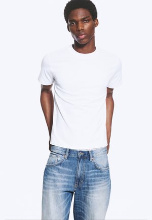Junger Mann, der ein einfarbiges weißes T-Shirt und hellblaue ausgewaschene Jeans trägt, steht mit den Händen hinter dem Rücken vor einem weißen Hintergrund.
