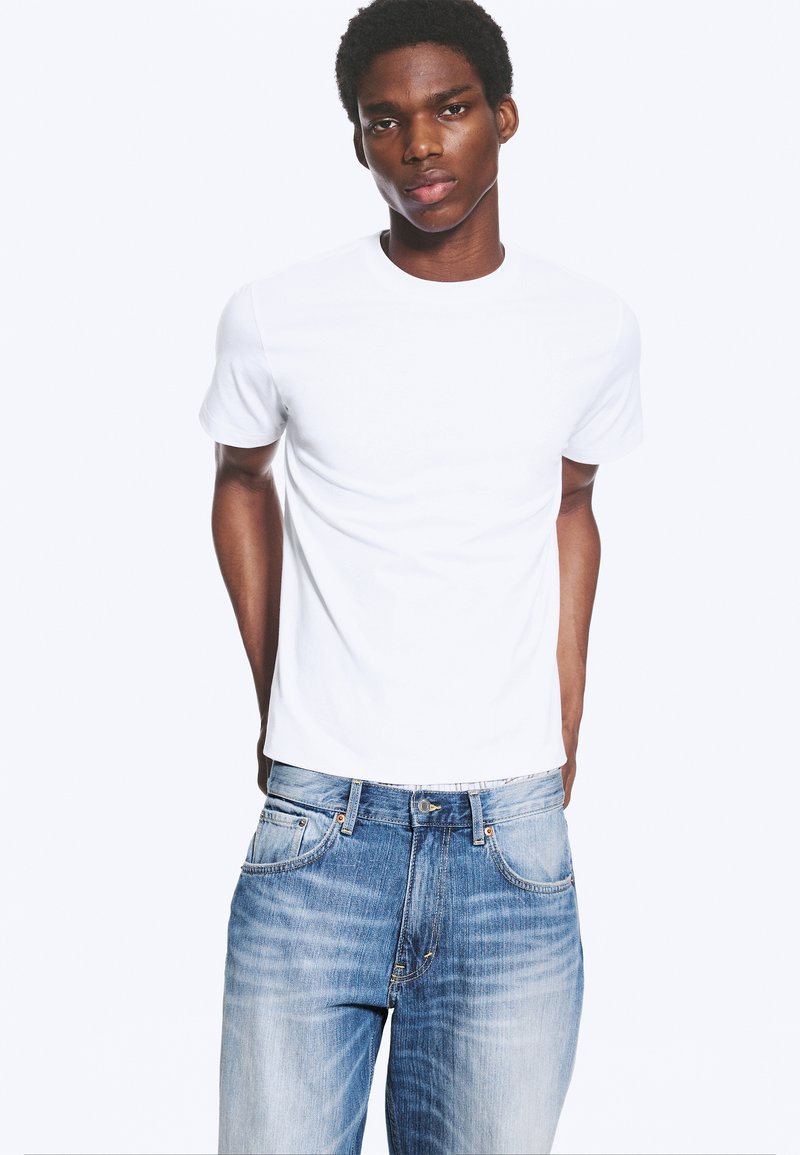 Jeune homme portant un t-shirt blanc uni et un jean bleu clair délavé, debout les mains derrière le dos sur un fond blanc.