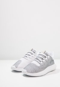 Vita atletiska skor med en texturerad grå stickad ovandel, rund tå och vit sula. Har snörningsdesign och vadderad krage.