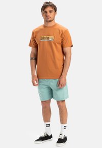 Camiseta de algodón naranja de manga corta con el logo de Quiksilver. Combinada con pantalones cortos azul claro, zapatillas negras y calcetines blancos con acentos de logo.