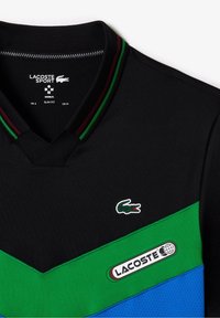 Svart Lacoste Sport skjorta i slim fit med gröna och blå chevronpaneler, krage i grön och burgundy rand, och krokodil-logotyp på bröstet.