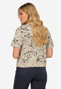 Camisa de manga corta beige con un patrón de dibujo de un pez en negro en la parte posterior. Combinada con pantalones azul marino, mostrando un estilo casual.