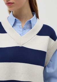 Gilet lavorato a maglia a righe blu e bianche con scollatura a V e bordi a coste, indossato sopra una camicia button-up azzurra chiara. Texture morbida.