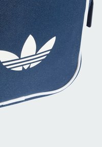 Plava platnena torba s bijelim Adidas logotipom koji sadrži tri listasta ukrasa i tri vodoravne pruge. Završena s bijelim obrubom.