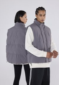 Pilkas pūkinis liemenė su aukštu apykakle, užtrauktuko uždarymu ir dviem priekinėmis kišenėmis. Siūlo elegantišką, siuvinėtą dizainą ir glotnų paviršių.