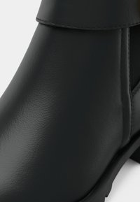 Gros plan d'une botte en cuir noir montrant une texture lisse, des détails de couture et une partie de la semelle sur fond blanc.