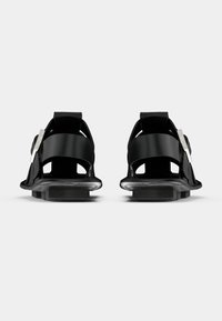 Τα μαύρα σανδάλια διαθέτουν μια ρευστή σχεδίαση με φαρδιές λουρίδες, γωνιώδη σόλα και μεταλλικές λεπτομέρειες, παρουσιάζοντας μια μοντέρνα, μινιμαλιστική αισθητική.