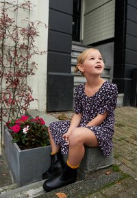 Čierne gumové čižmy so žltými akcentmi, čierne šaty s kvetinovým vzorom a nafúknutými rukávmi, vlasy v drdole, sediaca na kamennom schodíku pri kvetoch.