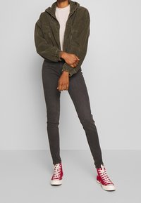 Olijf groene ribfluwelen zip-up jas met een capuchon, gecombineerd met een donkere spijkerbroek en rode hoge sneakers. Eenvoudig, gestructureerd ontwerp met voorzakken.