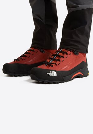 Rode en zwarte wandelschoenen met gestructureerd materiaal, een opvallend vetersysteem en een groot wit logo aan de zijkant. Stevige rubberen zool voor grip.