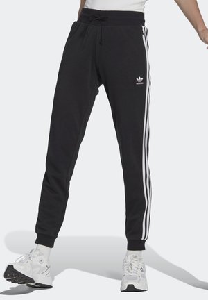 Pantalones deportivos Adidas negros con rayas laterales blancas y logo, usados con zapatillas blancas y una camisa blanca, mostrados desde la cintura hasta los pies.