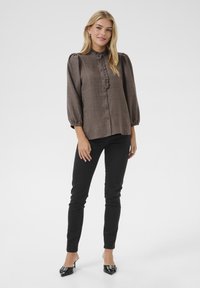 Chemise marron à manches bouffantes et détails en volants, assortie à un pantalon noir ajusté et des mules noires à talons. Texture du tissu lisse.