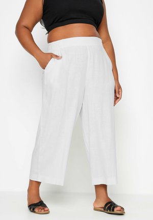 Person wearing loose white cropped pants with pockets, black crop top, and black slide sandals standing against a white background.