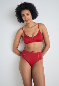 Crveni bikini set koji se sastoji od bralet vrha s izrezom u obliku žlice i visokog donjeg dijela s metalnim gumbima sa strane. Glatka tekstura.