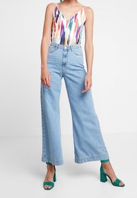 Kolorowy, wzorzysty kombinezon z szerokimi ramiączkami; zestawiony z jasnoniebieskimi, wysokim stanem, szerokimi spodniami typu jeans. Zielone sandały na obcasie dopełniają look.