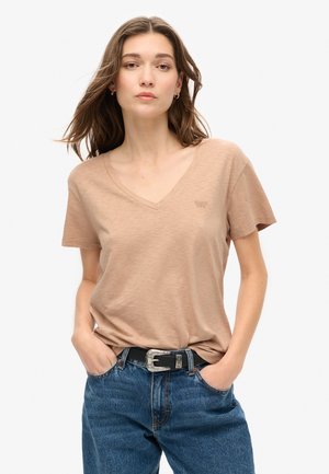 Kvinde iført beige T-shirt med V-hals, stukket ned i blå jeans med sort bælte, stående med hænderne i lommerne mod en ensfarvet baggrund.