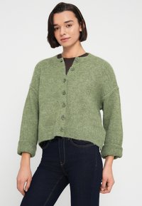 Πράσινη πλεκτή ζακέτα με γιακά λαιμόκοψη, κλείσιμο με έξι κουμπιά, κατεβασμένους ώμους και ρολαριστά μανίκια. Ριμπ τελείωμα και απαλό ύφασμα.