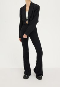 Person som bär en kort svart kavaj, vit crop top, ribbade utsvängda svarta byxor och svarta skor med tjock sula mot en vit bakgrund.