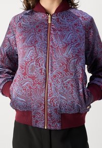 Chaqueta bomber en burdeos con patrón de cachemira azul, cuello, puños y cintura de canalé; presenta una cremallera dorada y bolsillos laterales.
