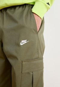 Λαδί cargo παντελόνια με πλαϊνές τσέπες, που φέρουν το λευκό λογότυπο της Nike. Το ύφασμα φαίνεται λείο και ελαφρύ, με άνετη εφαρμογή.