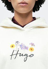 Fehér kapucnis pulóver virágmintás grafikával, melyen sárga és lila virágok láthatók, középen pedig fekete írott betűkkel a "Hugo" felirat szerepel.