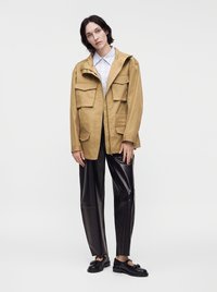 Femme portant une veste beige à grandes poches, une chemise blanche à col, un pantalon en cuir noir et des mocassins noirs, debout devant un fond blanc.