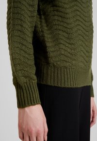 Maglione lavorato a maglia verde oliva con un motivo a onde testurizzato, dotato di polsini e orlo a coste. Il tessuto sembra morbido e leggermente spesso.