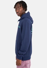 Sudadera con capucha azul navy, hecha de una mezcla de algodón, que cuenta con un bolsillo frontal tipo canguro, capucha con cordón y texto gráfico verde en el pecho y la manga.