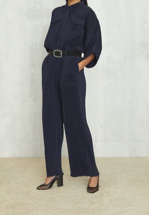 Kobieta ubrana w granatowy plisowany kombinezon z szerokimi rękawami, czarny pasek z ozdobną klamrą oraz czarne siateczkowe buty na wysokim obcasie, stojąca.