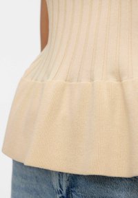 Beige geripptes Stricktop mit einem ausgestellten Saum, das vertikale Streifen und eine weiche Textur aufweist, kombiniert mit blauen Denimhosen.