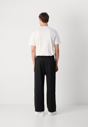 Mann, der mit dem Rücken zur Kamera steht, trägt ein weißes kurzärmeliges Hemd, schwarze lockere Hose und weiße Schuhe vor einem schlichten hellen Hintergrund.