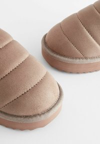 Des chaussures slip-on en daim brun avec un design matelassé, une texture douce et une semelle en caoutchouc présentant un motif ondulé et un bord coordonné.
