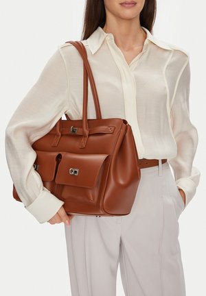 Kobieta ubrana w kremową przezroczystą bluzkę i jasne spodnie, trzymająca dużą brązową skórzaną torebkę z przednimi kieszeniami i srebrnymi zatrzaskami.