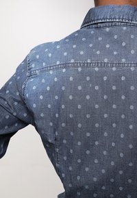 Denimskjorta med knapp-down krage, mörkblått tyg och ett ljusblått prickigt mönster. Synliga sömmar längs sömmarna och axlarna.