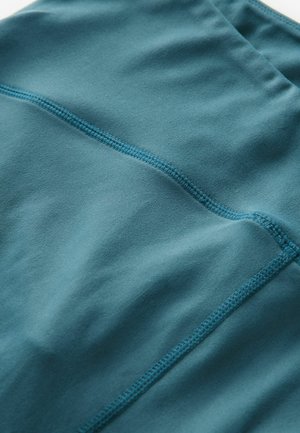 Tyrkysová sportovní látka s plochými švy, hladkým povrchem a mírným leskem. Vyznačuje se minimalistickým designem a přiléhavým tvarem.