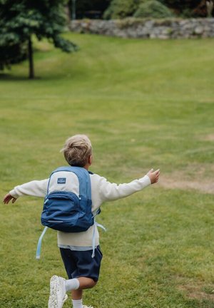 Bambino con capelli biondi che indossa un maglione bianco e uno zaino blu corre a braccia aperte su un prato nel parco.