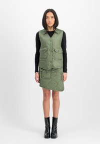 Grön kviltad väst med två framfickor, lager på lager över en matchande kjol. Outfiten har rundade former och knappdetaljer. Svarta ankelboots.