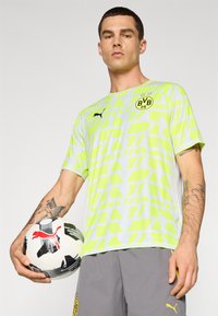 Camiseta de fútbol de manga corta con un patrón geométrico en verde y amarillo con detalles en negro, llevada por una persona que sostiene un balón de fútbol en blanco y negro.