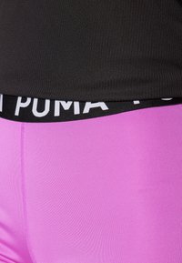 PUMA atletske legice s širokim črnim pasom, ki vsebuje belo blagovno oznako; svetlo roza tkanina z gladko, elastično teksturo.