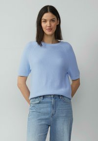Pull en maille bleu clair à manches courtes et col rond, avec des poignets côtelés, porté avec un jean en denim clair taille haute.