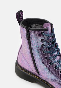 Botín brillante púrpura con un empeine de malla texturizada, cremallera negra y cordones negros. Presenta un detalle de costura amarilla alrededor de la suela.