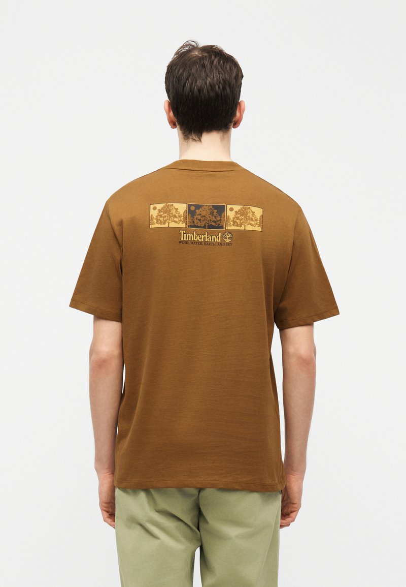 Άνδρας που φορά καφέ μπλούζα Timberland με τρία δέντρα και το κείμενο "Άνεμος, Νερό, Γη και Ουρανός" στο πάνω μέρος της πλάτης, συνδυασμένη με ανοιχτό πράσινο παντελόνι.