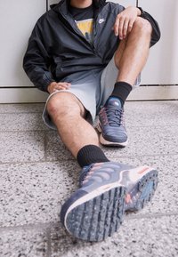 Nike-kengät harmaana ja sinisenä, tekstuurilla varustettuna, vaaleanpunaisilla yksityiskohdilla ja kestävä pohja, yhdistettynä mustiin sukkiin ja vaaleisiin shortseihin.