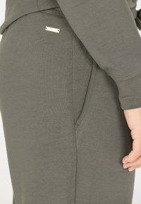 Olijfgroen stof met een zachte textuur, voorzien van een zijzak en een klein metalen accentlabel. Ontwerp met een ontspannen pasvorm en opgeslagen mouwen.