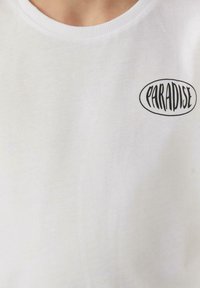Witte katoenen t-shirt met een ronde halslijn en zwarte "PARADISE" tekst in een ovale vorm aan de linkerkant. Glad materiaal, casual pasvorm.