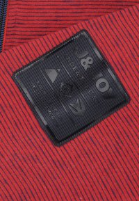 Roodgestreept stof met een gestructureerd oppervlak, voorzien van een zwartleren label. Het label toont logo's en tekst in wit en grijs tegen een geribbeld achtergrond.