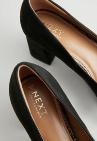 Zapatos de tacón alto de ante negro con un tacón cuadrado. El interior presenta un forro beige y el logo dorado de "NEXT". Textura suave, diseño de punta redondeada.