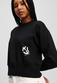 Μαύρο cropped sweatshirt φτιαγμένο από μαλακό υλικό, με λευκό λογότυπο στο στήθος. Μακριά μανίκια με απλό, καθαρό σχεδιασμό.