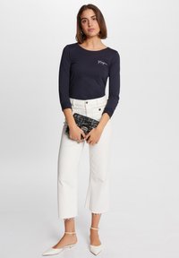Marineblaues Langarmoberteil mit weißem besticktem Logo, kombiniert mit hoch taillierten weißen Cropped-Hosen und einer schwarzen gemusterten Clutch. Weiße spitze Ballerinas.