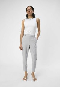 Pantalon tailleur gris clair avec une coupe slim et un détail de pli, associé à un top sans manches blanc et des tongs blanches. Texture de tissu lisse.
